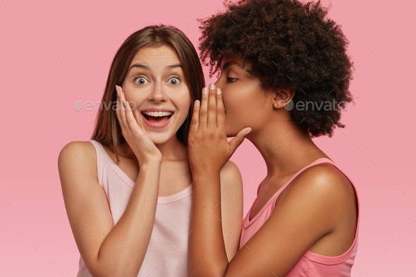 Young black woman whispers secret to her Caucasian friend, gossip together and spread rumours. Emoti