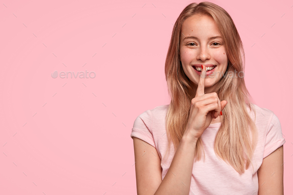 Pretty young female with cheerful expression, tells her secret to close friend, makes silence sign,