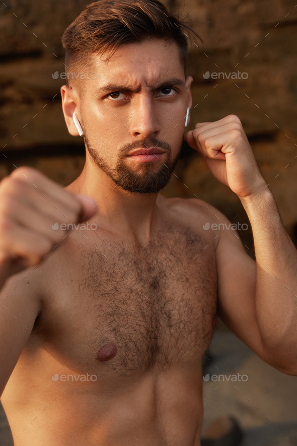Boxing man exercises in open air, has angry facial expression, keeps hands clenched in fists, looks