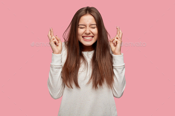 Pleased cheerful brunette woman puts all efforts in wishing good luck, crosses fingers, has faith fo