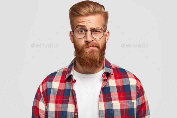 Photo of bewildered puzzled man with thick ginger beard and mustache, raises eyebrows, looks doubtfu