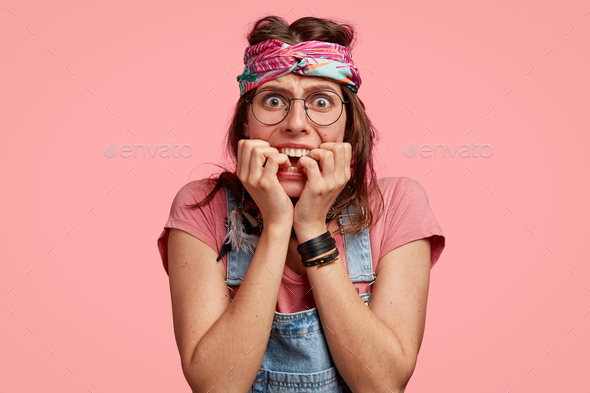 Photo of nervous anxious hippy woman bites finger nails, wears headband, round spectacles, dressed i