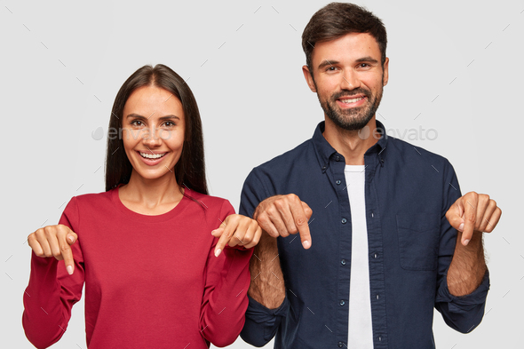 Horizontal shot of cheerful Caucasian young adult woman and man point with index fingers down, satis