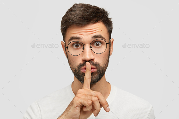 Unshaven Caucasian male makes silence gesture, asks to be quiet as someone sleeps, wears spectacles,