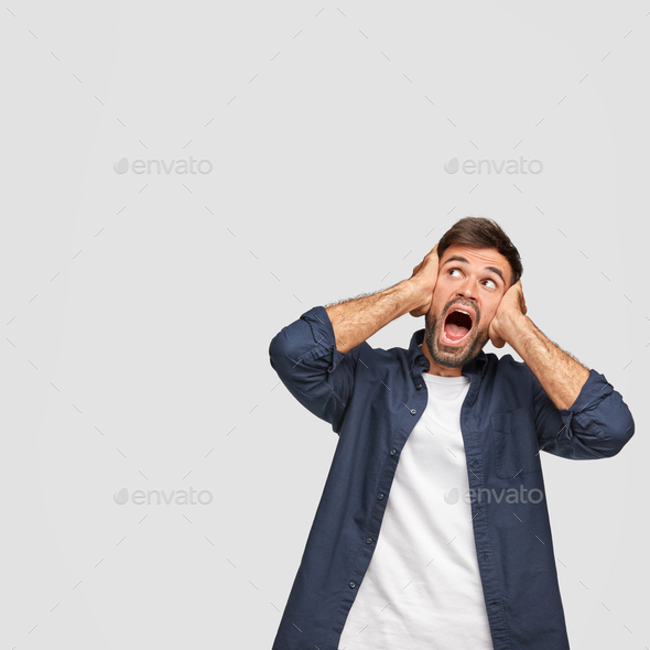 Vertical shot of stupefied Caucasian young man covers ears, opens mouth widely, sees something unbel