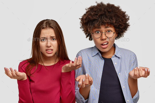 Photo of clueless two young multiethnic women with uncertain displeased looks, frown faces, spread p