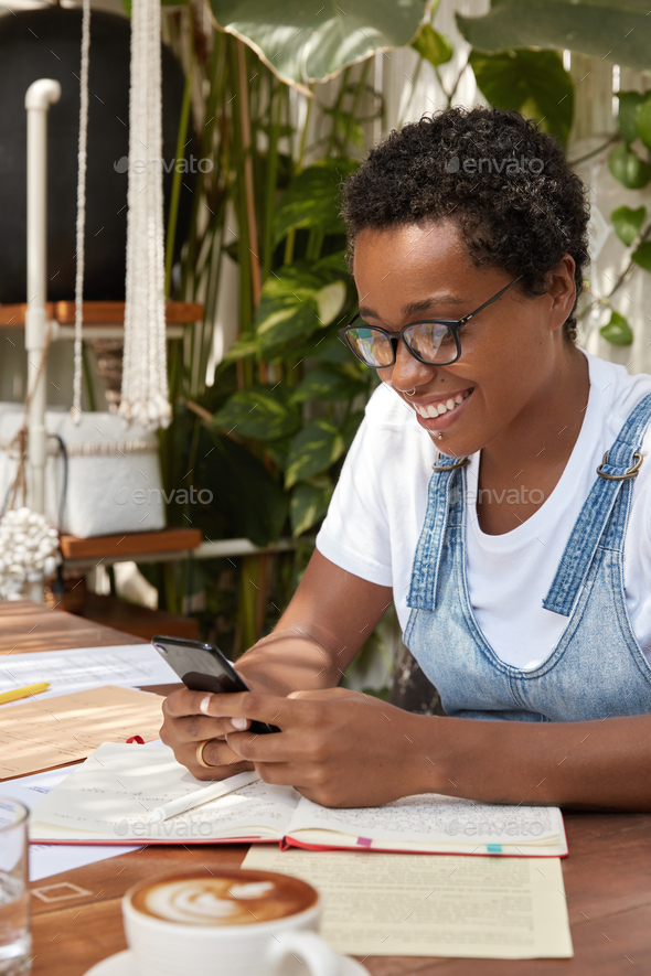 Photo of happy African American teenager glad to chat with friends on modern cellphone, has break af
