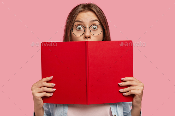Photo of intelligent female student covers face with red book, stares at camera with bugged eyes, fe