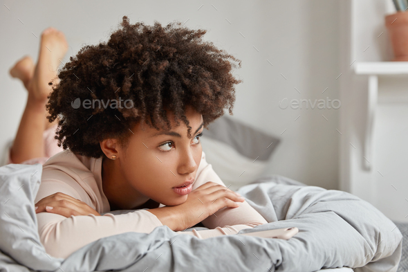 Cute African girl lies in bed, leans on hands, has thoughtful expression, enjoys comfort, being deep - Stock Photo - Images