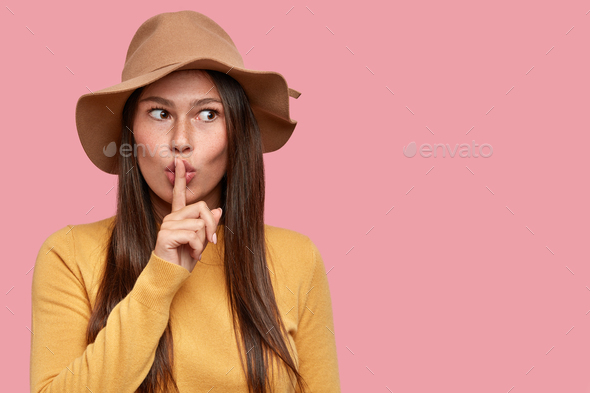 Brunette young woman with serious secret face, holds index finger in front of mouth, asks speak quie