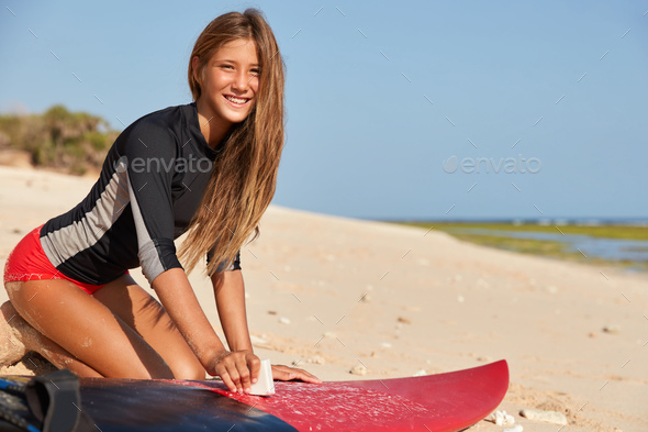 Happy young experienced surfboarder wears red bikini, has tanned skin, healthy body, waxes surfboard