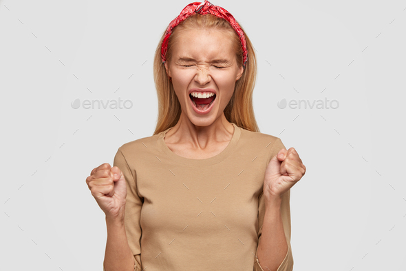 Photo of desperate blonde woman clenches fists, keeps mouth wide opened, expresses negativity, dress