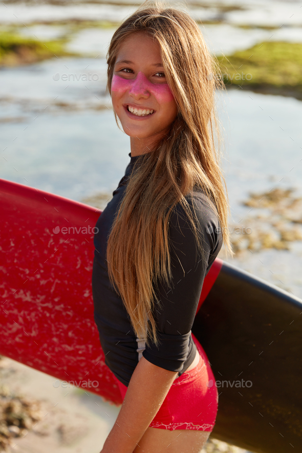Smiling active surfer has satisfied expression, long hair, protective sunscreen on face, wears water