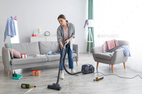 Unhappy young woman vacuuming messy apartment after party, cleaning up chaos after holiday
