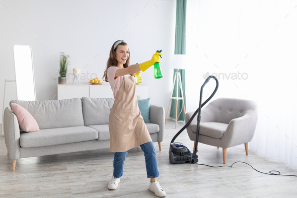 Happy professional maid with detergent spray bottles in middle of clean living room, copy space