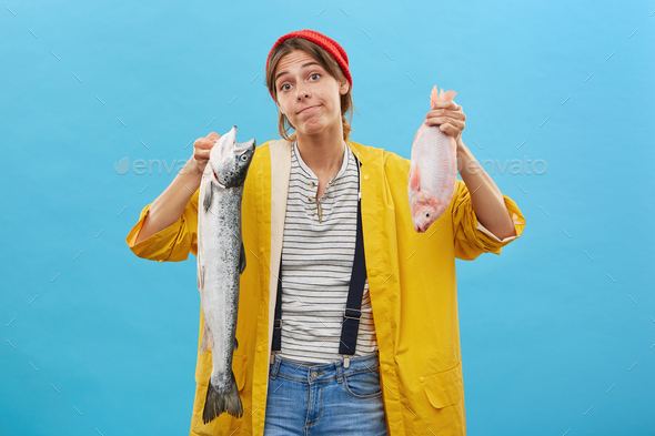 Young female angler looking with doubts at camera while holding two fish going to buy one of them, n