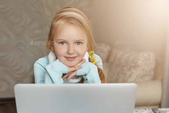 Adorable first-grade student girl with blonde hair in ponytail sitting in front of open laptop after