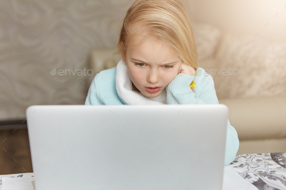 Stressed beautiful first-grade female pupil leaning on her elbow while doing homework using laptop p