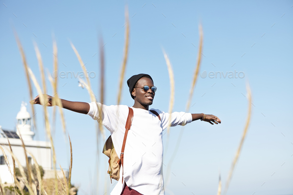 Cheerful stylish young Afro American tourist with knapsack wearing hat and stylish sunglasses spendi - Stock Photo - Images
