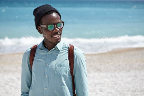 Cheerful young Afro American male student wearing eyeglasses and hat carrying knapsack on his should - Stock Photo - Images