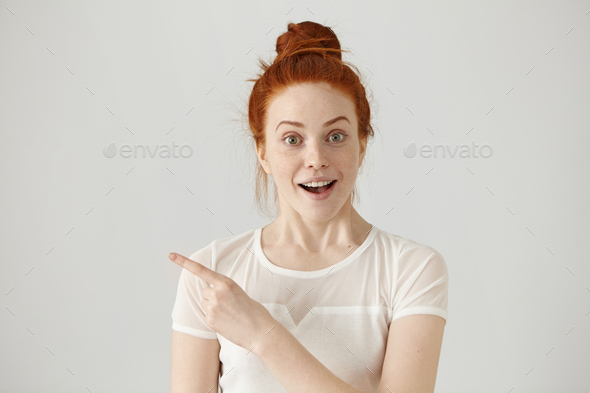 Excited young redhead Caucasian woman with hair knot pointing her index finger sideways, raising eye