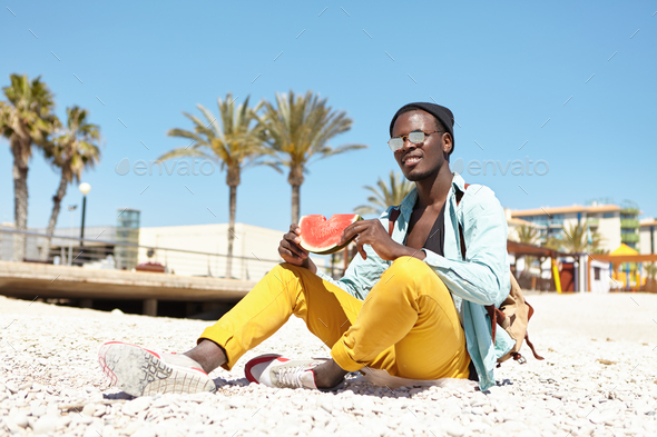 Outdoor portrait of handsome young African American man with knapsack sitting on pebble beach wearin - Stock Photo - Images