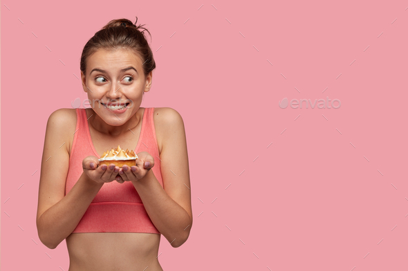 Photo of attractive joyful female bites lips, holds delicious cake, feels hungry after cardio traini