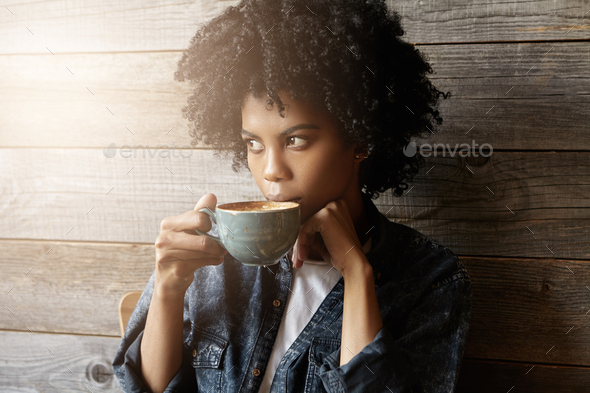 Indoor shot of beautiful African-American woman with Afro hairstyle holding big mug, having fresh ca