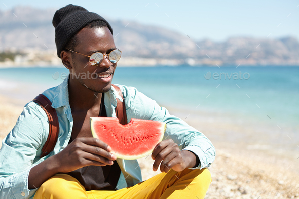Cheerful African American student wearing hat, mirror sunglasses and knapsack sitting cross legged o - Stock Photo - Images