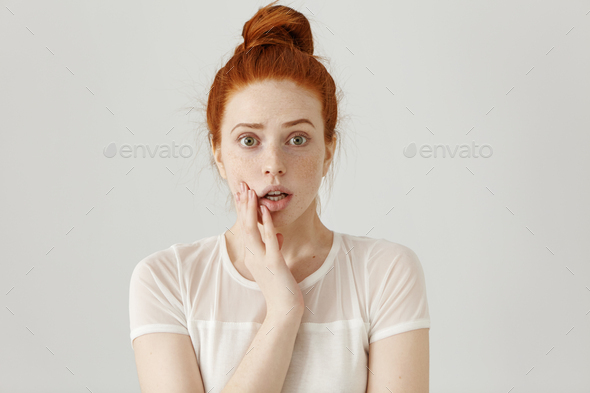 Frightened startled young redhead freckled female raising eyebrows, keeping hand on her cheek and st