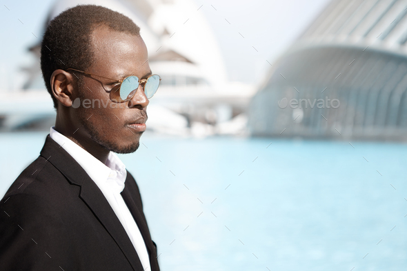 Profile of handsome successful young dark-skinned banker wearing round sunglasses and black suit sta