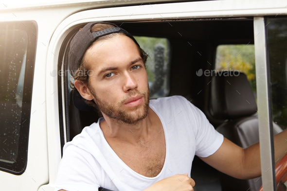 Close up view of handsome young man with stylish beard sitting on driver\'s seat in leather cabin of