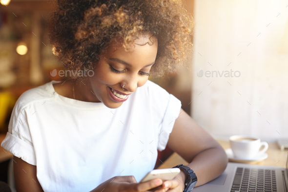 Adorable hipster dark-skinned woman with Afro hairstyle checking her news feed or messaging via soci