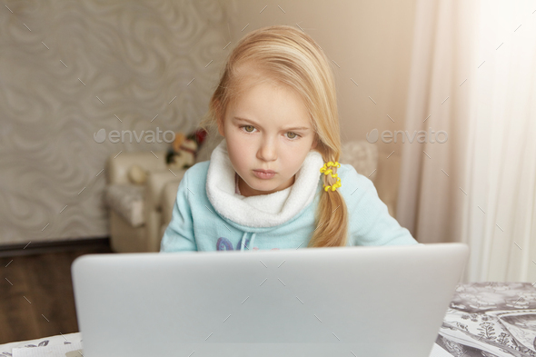 First-grade A-student girl with messy blonde hair sitting in her room in front of open laptop pc, lo