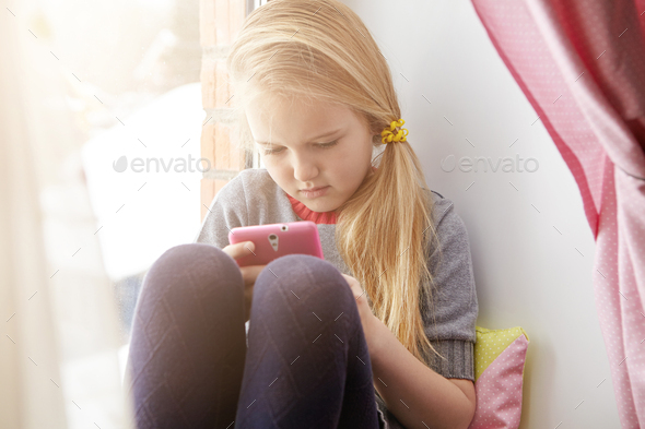 Serious concentrated adorable first-grade school girl sitting at window with cell phone, posting her