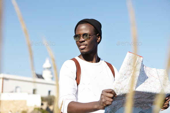 Summer portrait of handsome young black European man in trendy clothing using city guide while sight