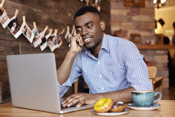 Indoor shot of attractive young dark-skinned employee in shirt using generic laptop computer, surfin
