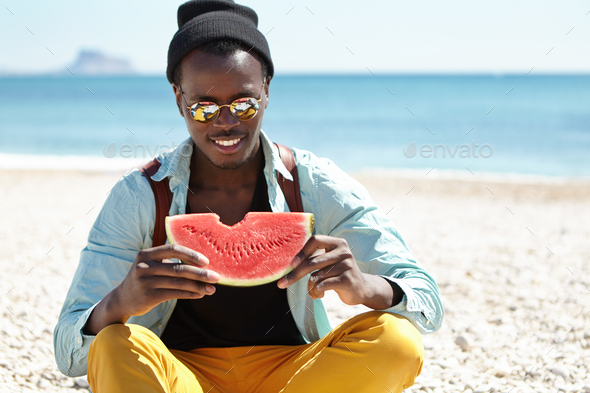 Trendy looking handsome young Afro American traveler with knapsack sitting on pebble beach against c - Stock Photo - Images