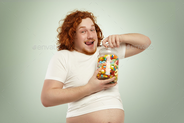 Portrait of happy excited obese young redhead male in undersized t-shirt opening jar of candies in h