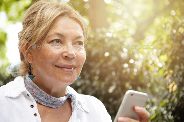 Close up shot of good-looking mature woman with fair hair wearing stylish clothes holding smart phon