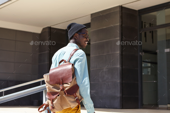 Rear shot of happy African American man traveler carrying leather knapsack about to enter modern hot - Stock Photo - Images