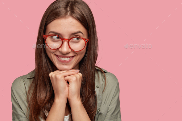 Headshot of dreamy pleased woman with tender smile, wears red rim spectacles, keeps hands under chin