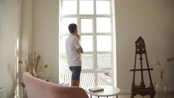 A man in a spacious room gets up from the sofa and goes to think to the big window alt