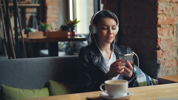 Young Woman in Headphones Listening To Music Using Smartphone in Modern Cafe alt