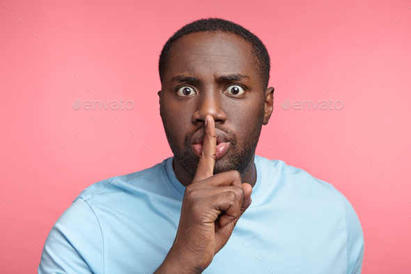 Buged eyed dark skinned male keeps fore finger on lips, being shocked as friends tells very private