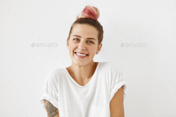 Glad blue-eyed female with pink hair knot, wearing loose white shirt, having tattooes on arms, smili