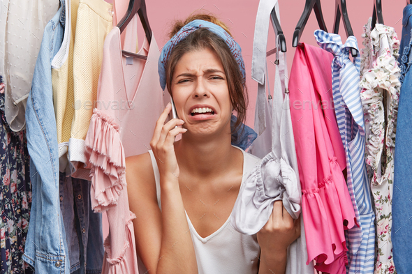 Upset woman standing near rack of clothes, chatting over smart phone with her friend, complaining th