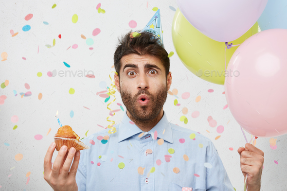Studio shot of surprised bearded male holds colorful balloons and cake with burning candle, anticipa