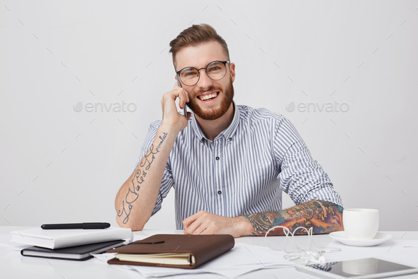 Candid shot of fashionable tattooed man wears shirt with rolled up sleeves and round glasses, calls