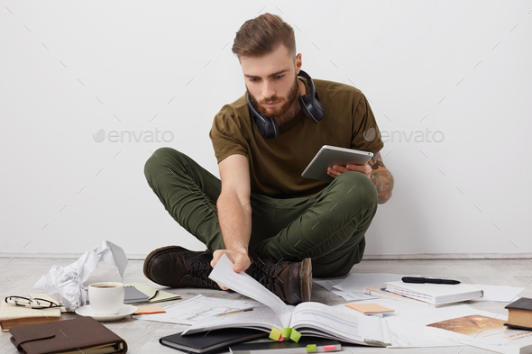 Preoccupied beared male college student with trendy hairdo looks attentively into book, holds modern